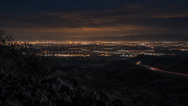 Henderson and the Las Vegas valley at night from a desert ridge: city lights in a wide band, a winding road with light trails below, low clouds lit orange