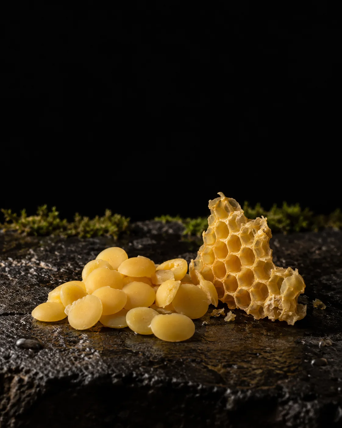 Golden beeswax pastilles beside a piece of wax honeycomb on wet black basalt, moss behind