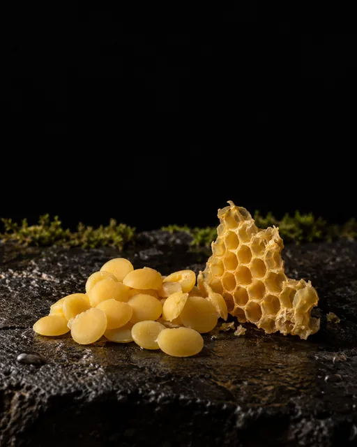 Golden beeswax pastilles beside a piece of wax honeycomb on wet black basalt, moss behind