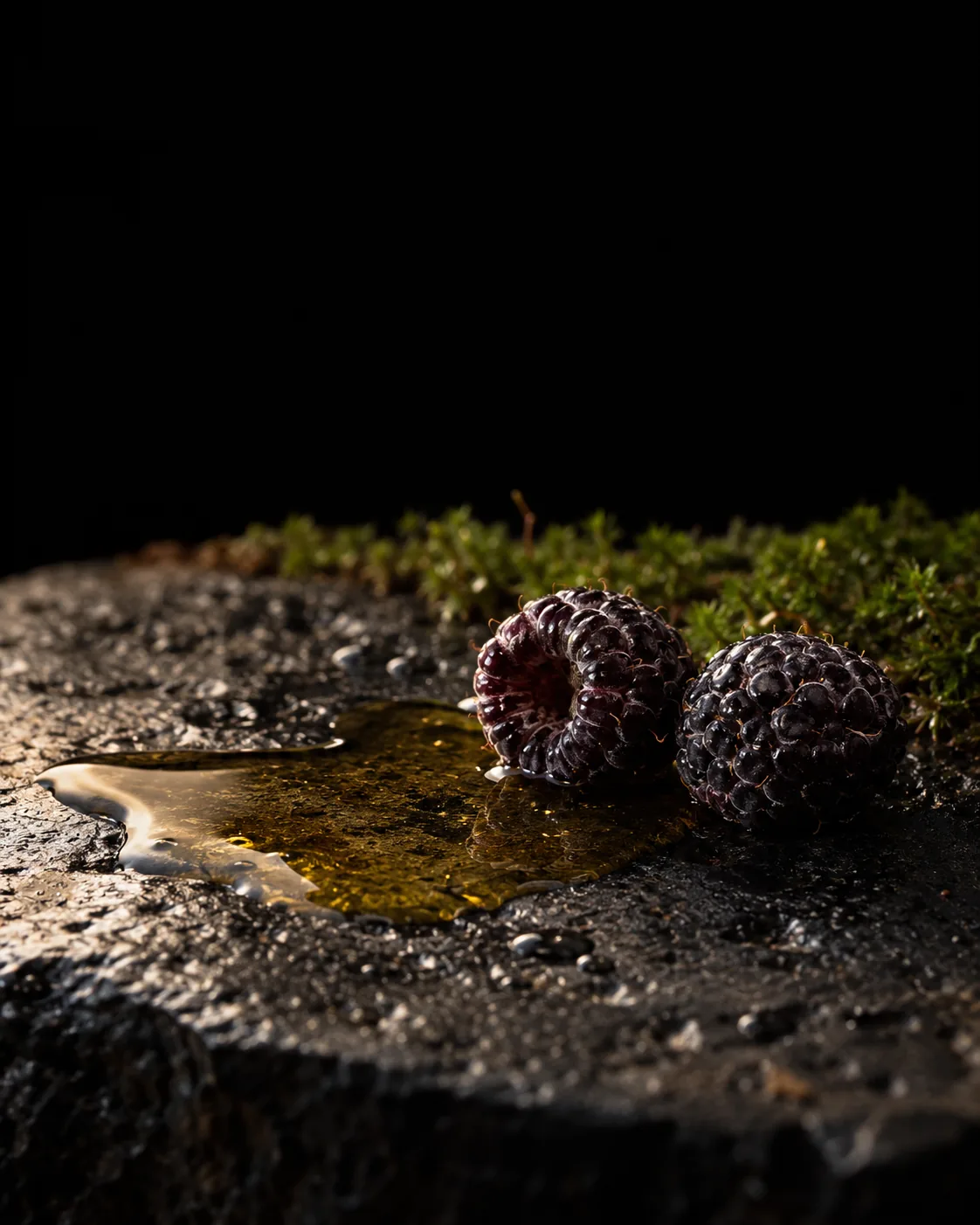 A pool of golden black raspberry seed oil beside two black raspberries on wet black basalt