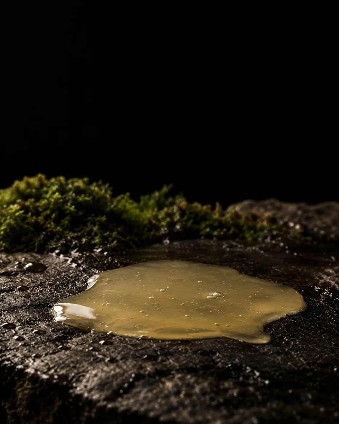 A small pool of hazy, pale yellow coco-glucoside liquid on wet black basalt, moss behind