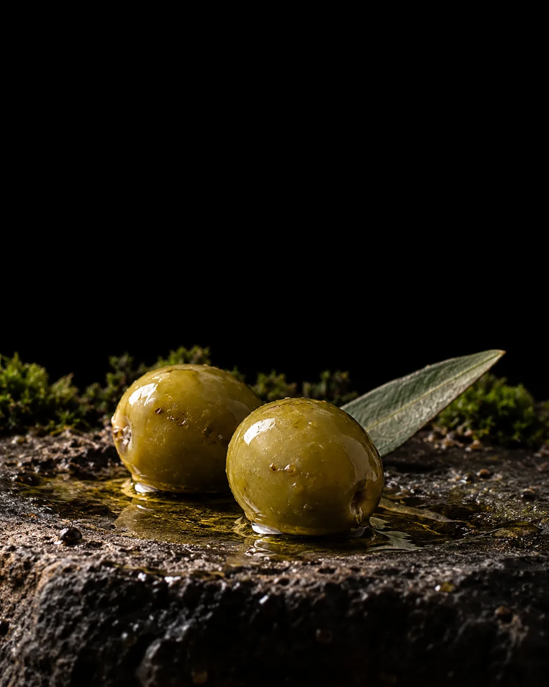 Two green olives and an olive leaf in a pool of golden-green olive oil on wet black basalt