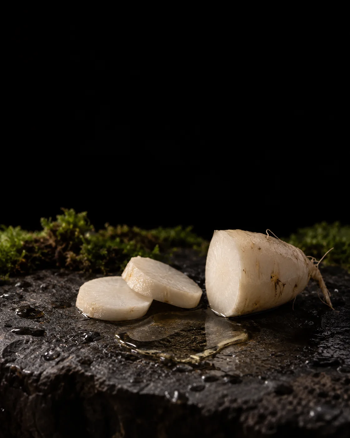 A small pool of pale ferment filtrate beside slices of fresh radish root on wet black basalt