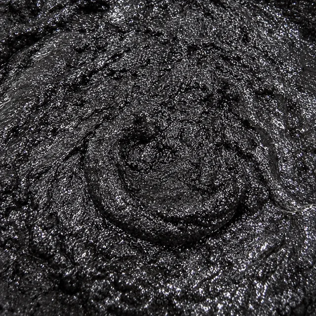 The glossy surface of jet-black sugar scrub with a spoon swirl through it: fine sugar soaked black with honey, glycerin and charcoal, glinting silver where the light hits