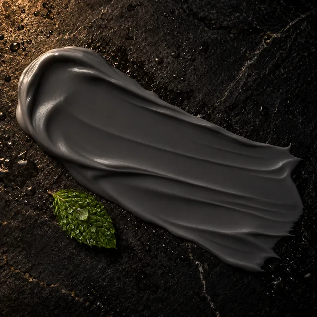 A smooth, thick swipe of charcoal-grey cream cleanser on dark wet stone, beside a peppermint leaf